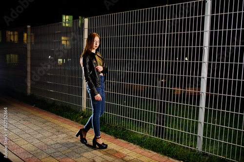 Night portrait of girl model wear on jeans and leather jacket against iron fence.