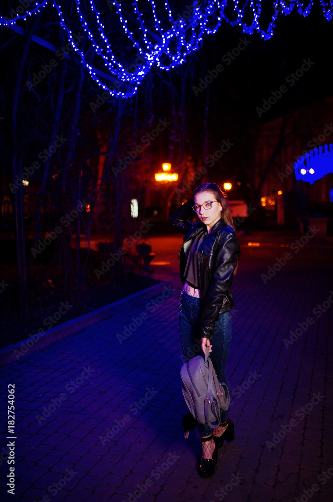 Night portrait of girl model wear on glasses, jeans and leather jacket, with backpack in hands, against blue lights garland of city street.