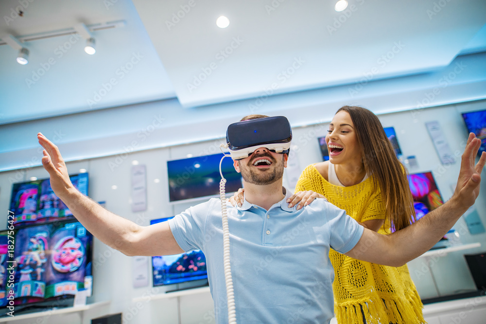 Fototapeta premium Satisfied young bearded handsome man tasting VR while his attractive smiling girlfriend standing behind him and holding neck with hands in a tech store.