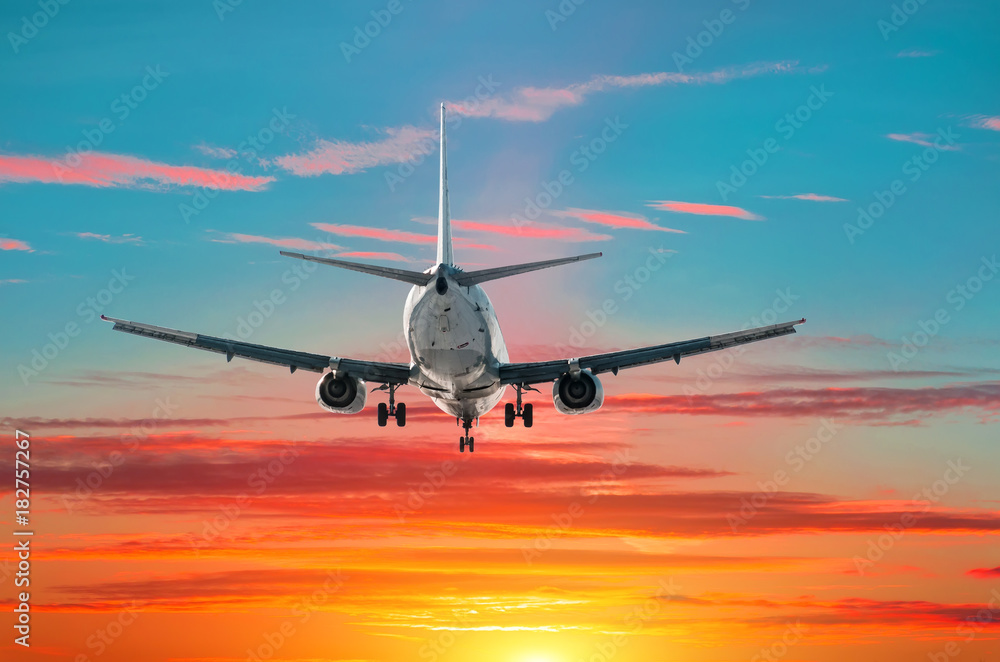 Passenger airplane flies landing at sunset on the background of blue green gradient and red sky.