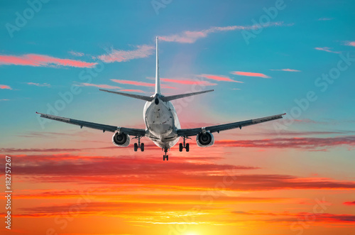 Passenger airplane flies landing at sunset on the background of blue green gradient and red sky.