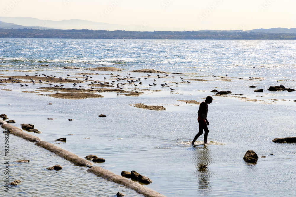 Φωτογραφία A man in swimsuit walks at the beach near Lago di Garda in Sirmione, Lombardy, n