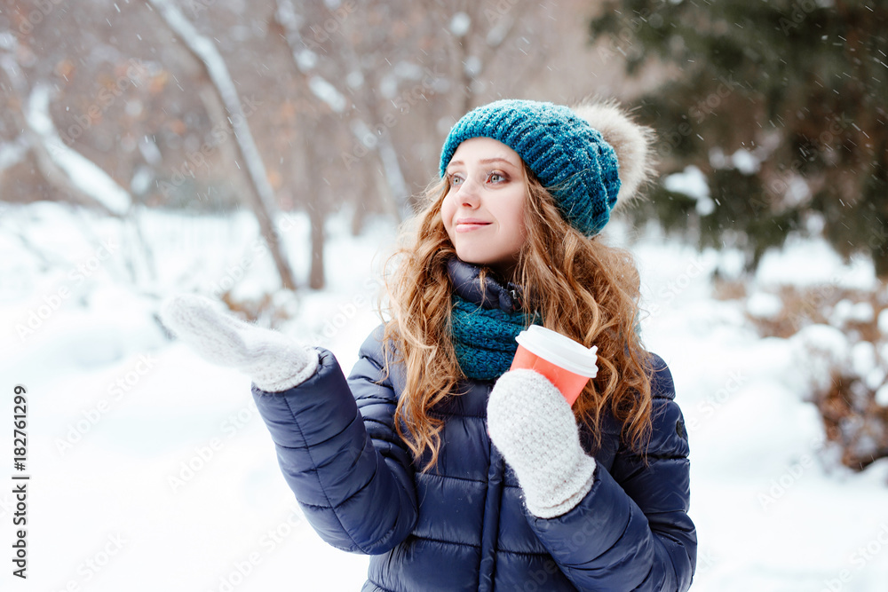 Winter walk in the park or the wood. The young beautiful girl the blonde with blue eyes in a warm cap and a down-padded coat catches a hand in a snowflake mitten. There is snowfall. Winter walk in the park or the wood. The young beautiful girl the blonde with blue eyes in a warm cap and a down-padded coat catches a hand in a snowflake mitten. There is snowfall.