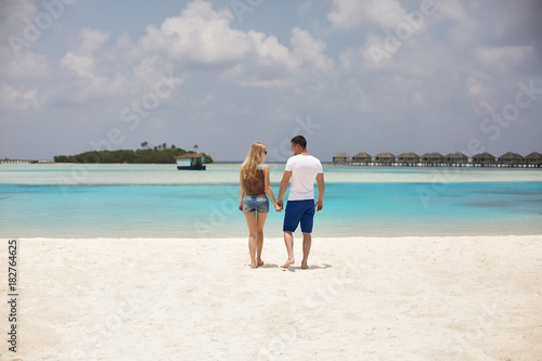 Back view of couple holding hands and walking to the blue ocean lagoon on Maldives at luxury spa resort. Travel and honeymoon concept.
