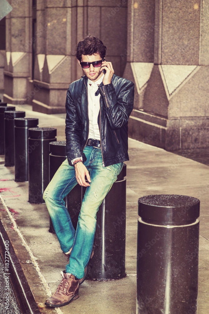 Dressing in black leather jacket, blue jeans, brown boot shoes, wearing  sunglasses, a young European Businessman with beard sitting on street in  New York, talking on mobile phone during working break. Stock