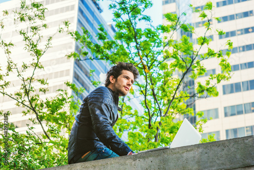 European graduate student studying in New York.  Young guy with beard, sitting on the top of wall on business district in spring day, working on laptop computer, confidently looking forward, thinking