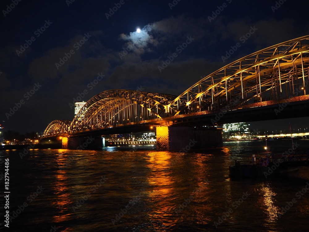 Fototapeta premium Deutzer Brücke in Köln bei Nacht mit verschwommenen Lichtern und Mond im Hintergrund