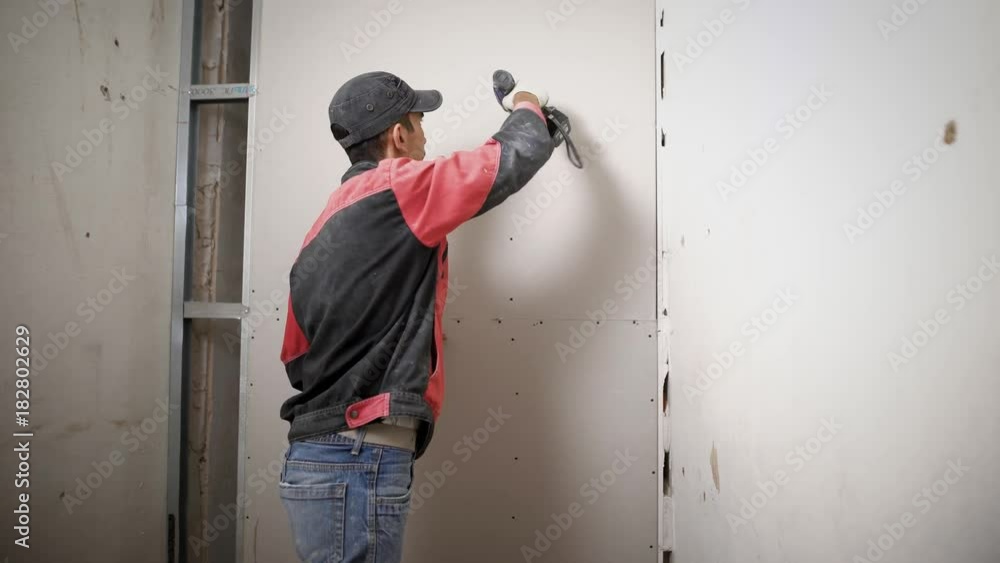 A professional builder screws the nails into the wall, which was made of gypsum, the man holds a drill in his hands and clogs the nails A professional builder screws the nails into the wall, which was made of gypsum, the man holds a drill in his hands and clogs the nails