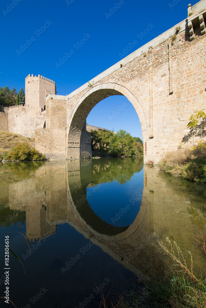 arch of Alcantara bridge, landmark and monument from ancient Roman age, reflected on water of river Tagus, Tajo in Spanish, in Toledo city, Spain, Europe
