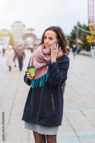 young beautiful brunette girl with coffee to go and smartphone on the background of blurred city street