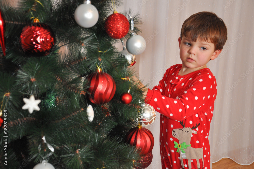 Boy in New Year's pajama decorating Christmas tree. Child putting decorations on Christmas tree