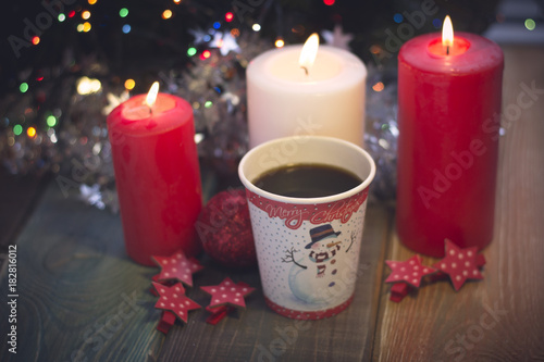 A romantic festive still life with burning candles and a paper cup of coffe with Christmas design. A colored wooden surface. White and red colors. Dark Christmas background. Blurred bokeh