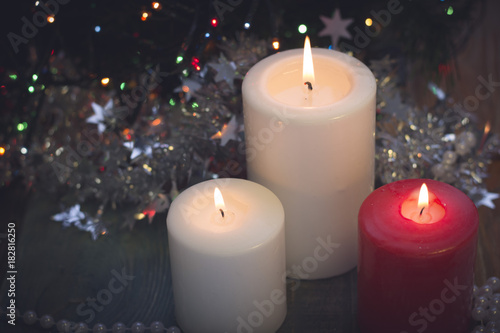 A romantic festive still life with burning candles of different size. White and red colors. Dark Christmas background. Blurred pine tree on the back. Blurred bokeh