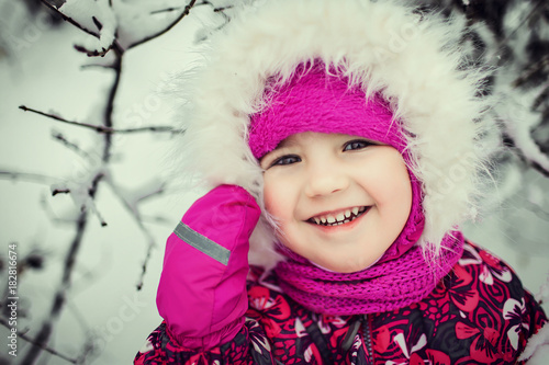 portraite of little cute kid in pink hat, hood with white fur and coat in pattern enjoying day out, smiling in the winter forest