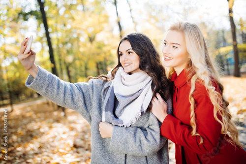 Two beautiful young woman taking selfie in autumn day in park