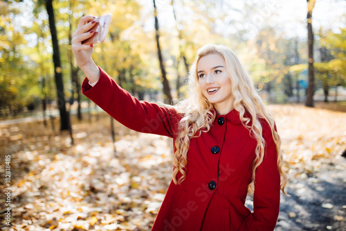Teenage girl in autumn in park taking a selfie on smart phone. Beautiful happy young woman taking a photo of herself outdoors on autumn day photo
