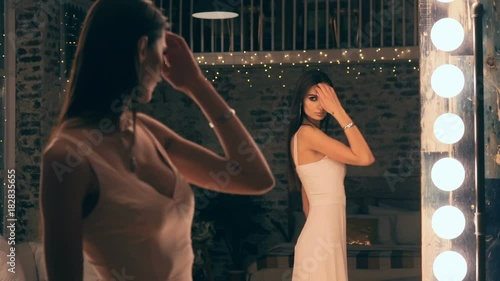 Portrait of elegant girl in evening dress posing in front of a mirror with the lights in the locker room. Brunette with makeup and hair.