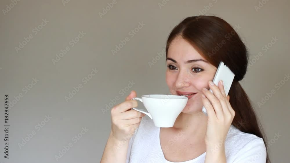 Portrait of a beautiful brunette girl who drinks a hot drink from a cup and talks on her mobile phone. Young woman is holding talks on a smartphone and smiling. Break Portrait of a beautiful brunette girl who drinks a hot drink from a cup and talks on her mobile phone. Young woman is holding talks on a smartphone and smiling. Break