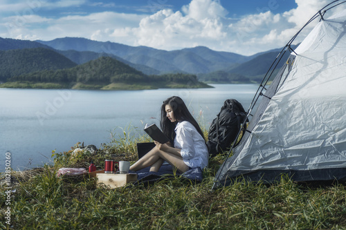 Pretty asian young woman reading book on tent camping nature near lake. 