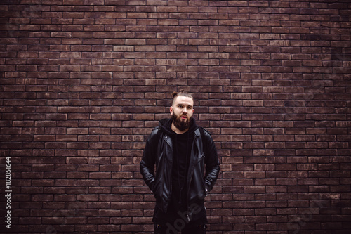 Modern young bearded man in black style clothes posing against brick wall.