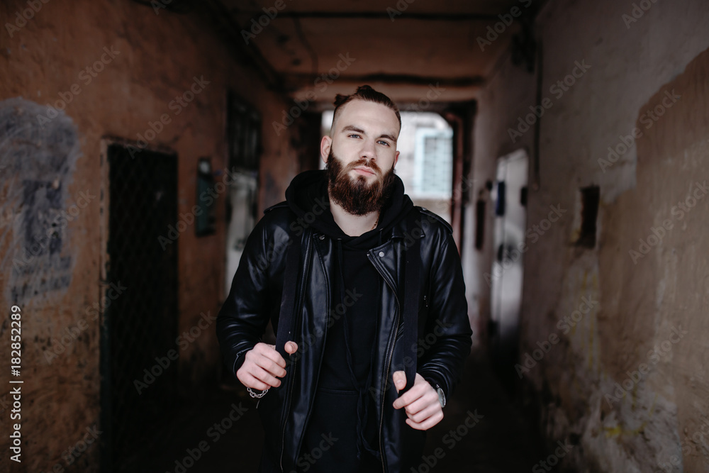 Modern young bearded man in black style clothes standing in around urban background.