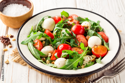 Fresh salad with arugula, cherry tomatoes, mozzarella cheese and walnuts on white wooden background.