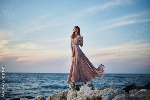 Beautiful young woman in a long dress standing by the sea, rocks