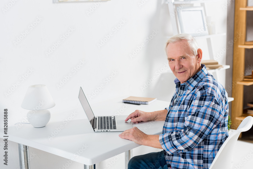 side view of senior man sitting at table with laptop and looking at camera at home side view of senior man sitting at table with laptop and looking at camera at home