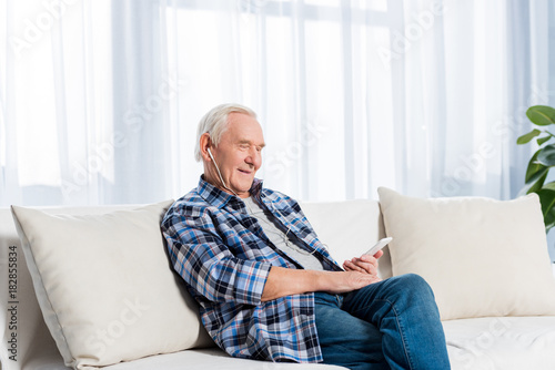 side view of smiling senior man in earphones with smartphone resting on sofa at home