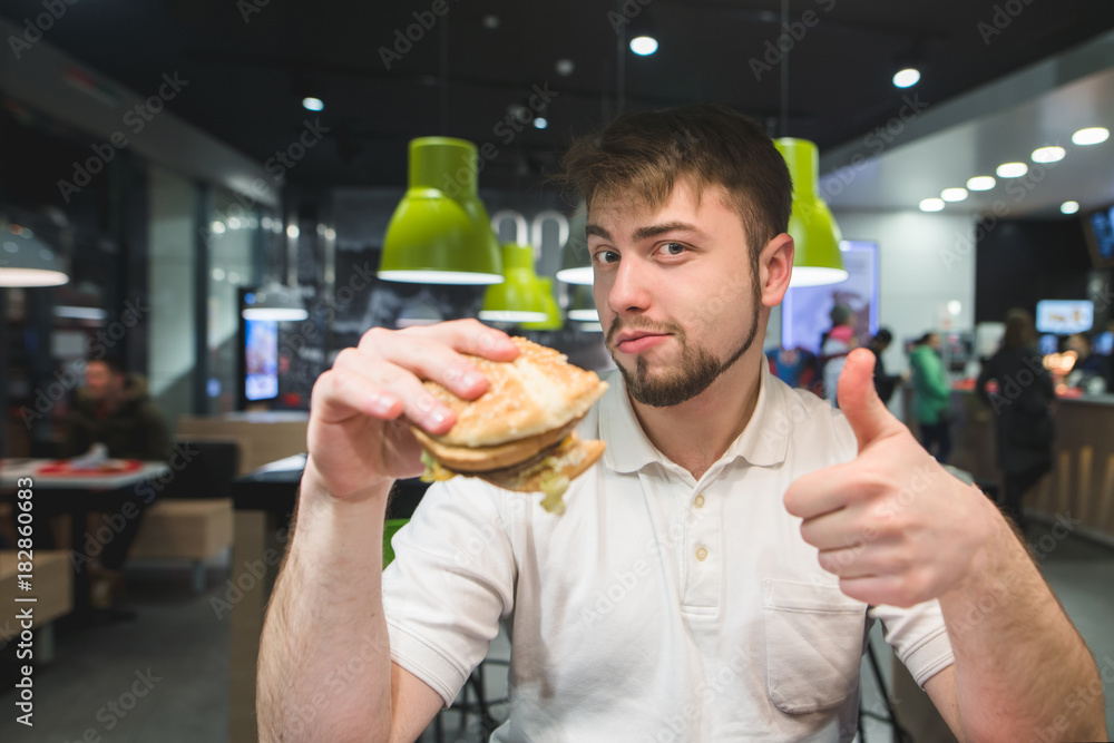 A man with a burger in his hands sits in a fast-food restaurant and shows a thumbs up. A man likes a burger. A man with a burger in his hands sits in a fast-food restaurant and shows a thumbs up. A man likes a burger.