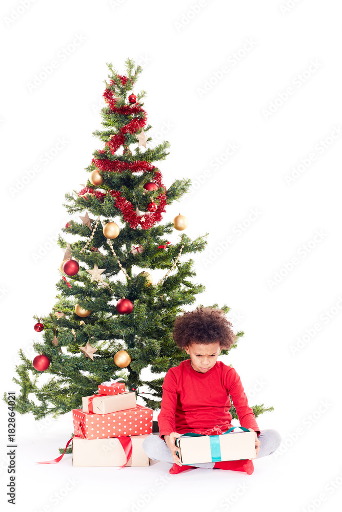 Portrait of cute mixed race little boy sitting on floor near Christmas tree with gift box in his hands Portrait of cute mixed race little boy sitting on floor near Christmas tree with gift box in his hands