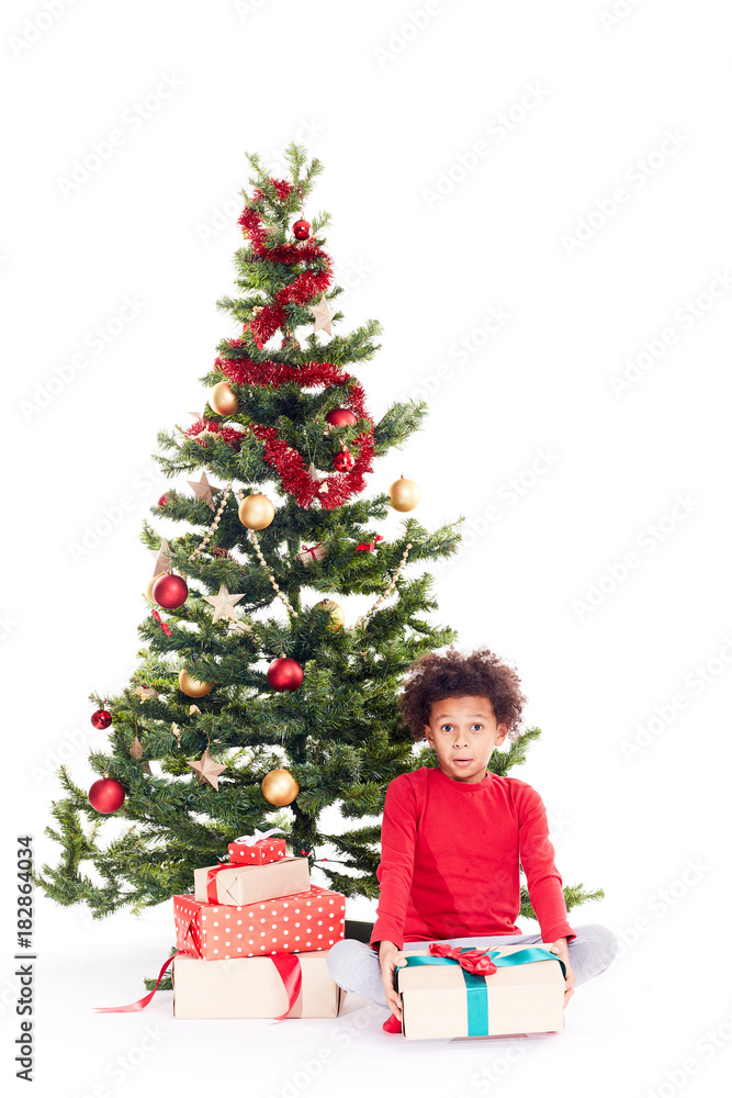 Portrait of cute mixed race little boy sitting on floor near Christmas tree with gift box in his hands Portrait of cute mixed race little boy sitting on floor near Christmas tree with gift box in his hands