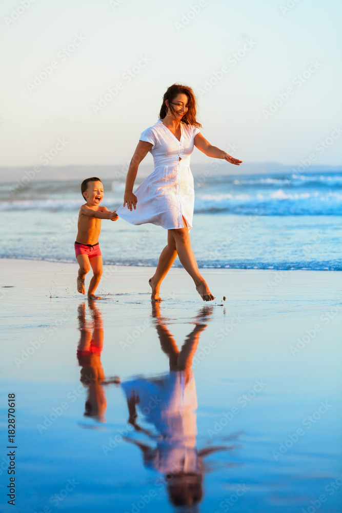 Happy family - mother, baby son have fun together, barefoot child run with splashes by water pool along sunset sea surf on black sand beach. Travel lifestyle, parents with kids on summer vacation.