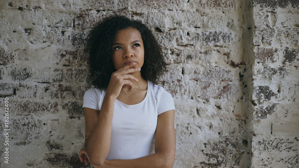 Thoughtful young mixed race girl thinking about solving problem on brick wall background Thoughtful young mixed race girl thinking about solving problem on brick wall background