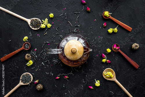 Brew the aromatic tea. Tea pot near wooden spoons with dried tea leaves, flowers and spices on black wooden background top view