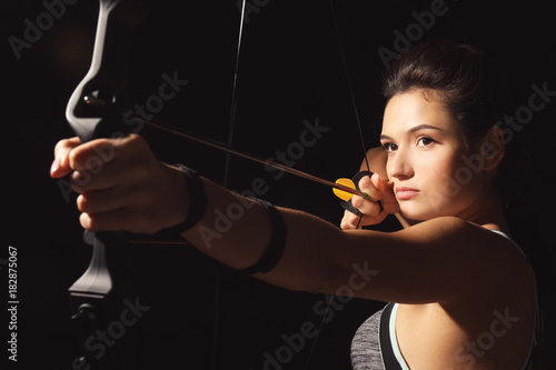 Sporty young woman practicing archery on dark background