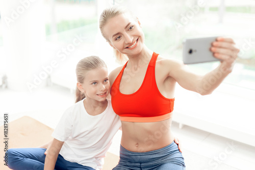 Mother and daughter are engaged in yoga in sportswear. They are in a bright room with panoramic windows.