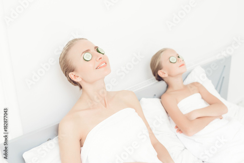 Mother and daughter had a day of spa. They are in white bath towels with slices of cucumber on their eyes