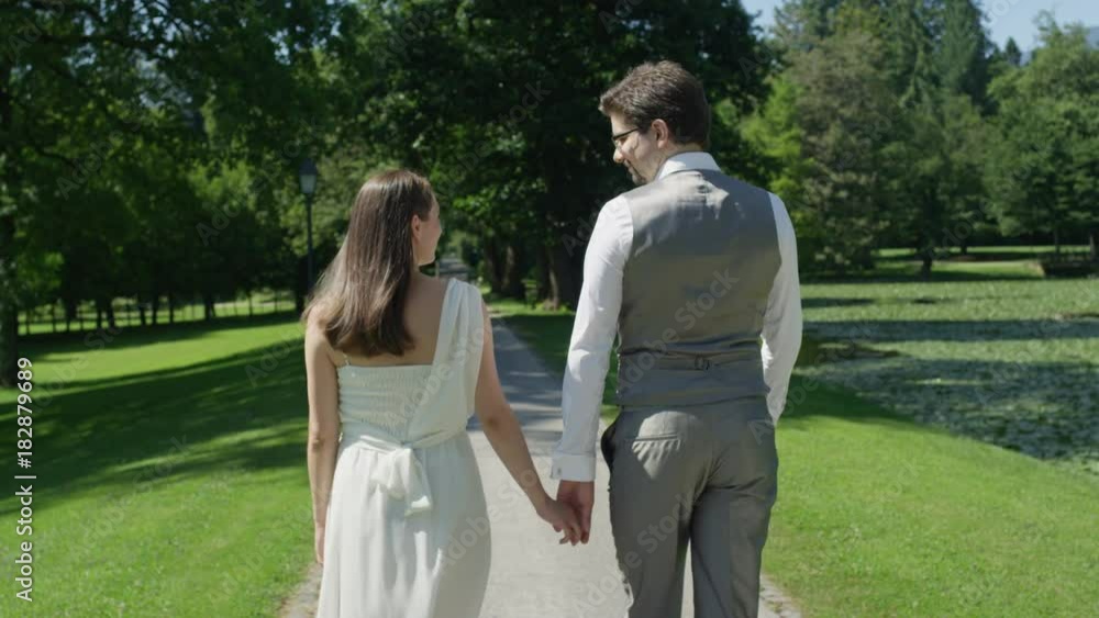 SLOW MOTION, BACK VIEW CLOSE UP: Happy married couple holding hands on a stroll through the park. Man and wife lovingly embrace walking on promenade in sunny spring. Young newlyweds expecting baby. SLOW MOTION, BACK VIEW CLOSE UP: Happy married couple holding hands on a stroll through the park. Man and wife lovingly embrace walking on promenade in sunny spring. Young newlyweds expecting baby.