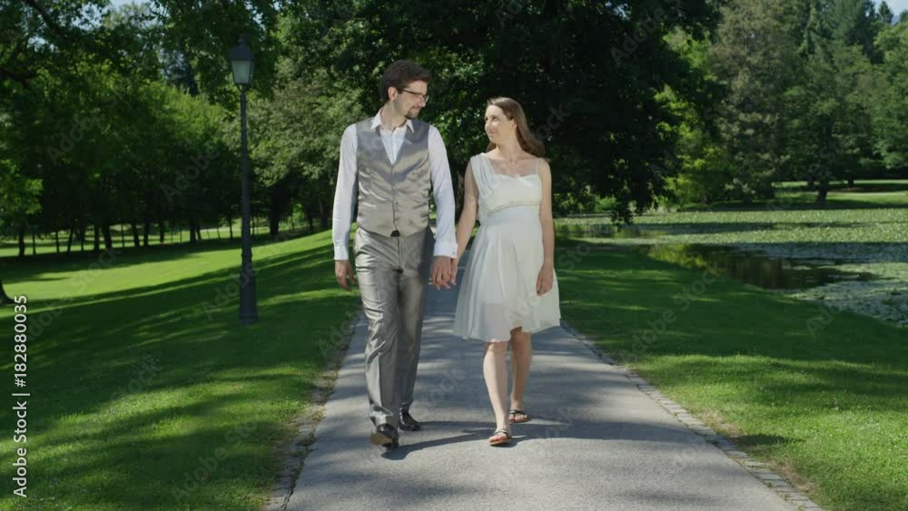 SLOW MOTION: Happy pregnant couple holding hands and walking trough green park in sunny spring. Loving man taking a stroll with his wife during pregnancy. Young newlyweds expecting baby after wedding SLOW MOTION: Happy pregnant couple holding hands and walking trough green park in sunny spring. Loving man taking a stroll with his wife during pregnancy. Young newlyweds expecting baby after wedding