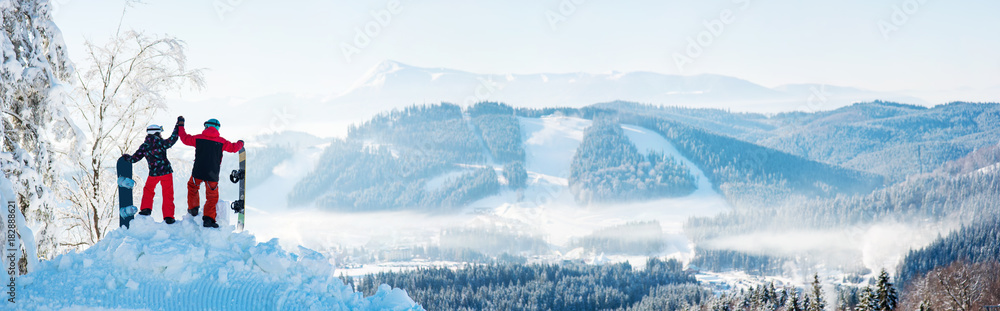 Winter panorama of the Carpathians mountains landscape and forests in a white haze. In the side couple high fiving each other on top of a snowy mountain, resting after snowboarding at ski resort - obrazy, fototapety, plakaty 