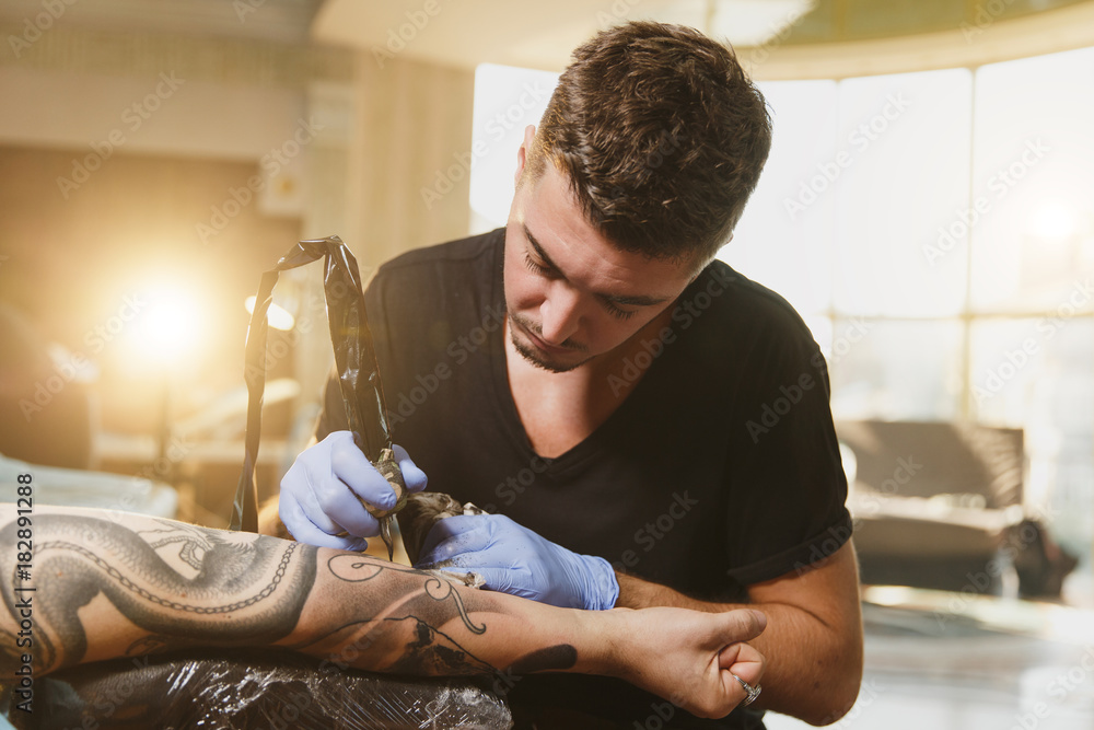 Professional tattooer artist doing picture on hand of man by machine black  ink from a jar. Tattoo art on body. Equipment for making tattoo art. Master  makes tattooed in light studio. Stock