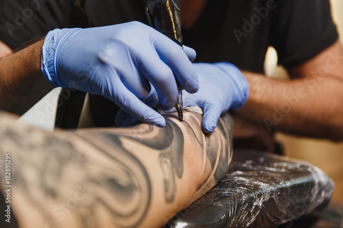 Close up of professional tattooer artist doing picture on hand of man by machine black ink from a jar. Tattoo art on body. Equipment for making tattoo art. Master makes tattooed in light studio. photo
