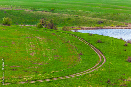 A rural road running along the river bank next to a plowed field. Green spring in Russia.