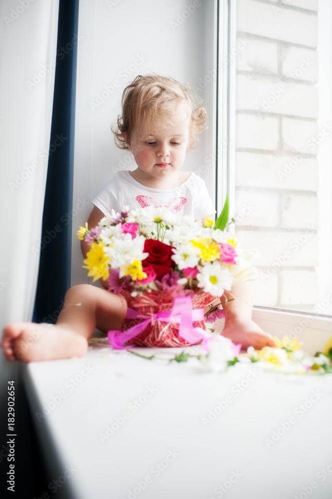 Baby sitting on a white window sill with a bouquet of flowers and looking at them Baby sitting on a white window sill with a bouquet of flowers and looking at them
