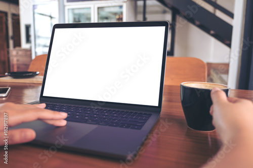 Mockup image of a hand using and touching laptop with blank white screen while drinking coffee on wooden table in cafe