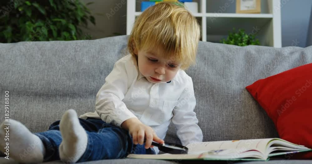 Cute small boy sitting on the couch and looking at the book page through the dandruff in his hand in the living room. Indoors Cute small boy sitting on the couch and looking at the book page through the dandruff in his hand in the living room. Indoors