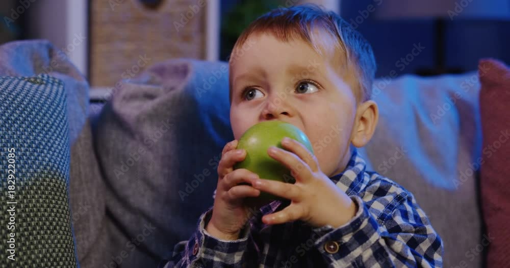 Close up of the funny small boy biting a big green apple and chewing it on the sofa with pillows at night. At home. Indoor. Portrait Close up of the funny small boy biting a big green apple and chewing it on the sofa with pillows at night. At home. Indoor. Portrait