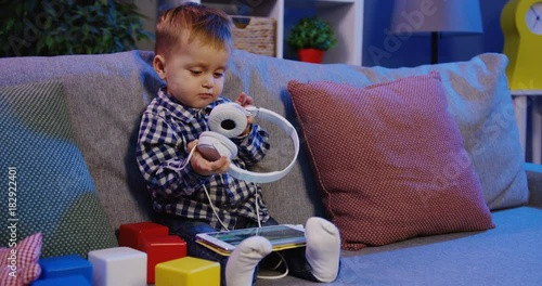 Little blonde nice boy taking on big white headphones and preaparing for listening to the music on the tablet while sitting on the sofa among kid's cubes and pillows. At home. Inside photo