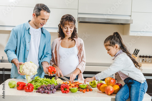 cheerful family with pregnant mother slicing vegetables and fruits at kitchen